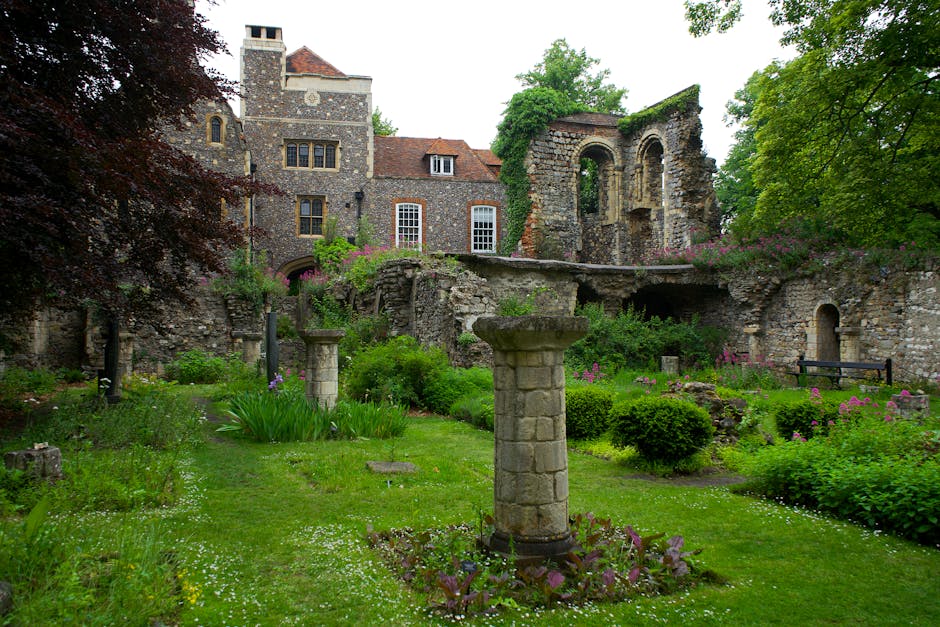 A scenic medieval-style garden featuring well-maintained green lawns and a variety of lush plants. In the foreground, there is a stone pedestal with a flat top, surrounded by flowerbeds with small pur