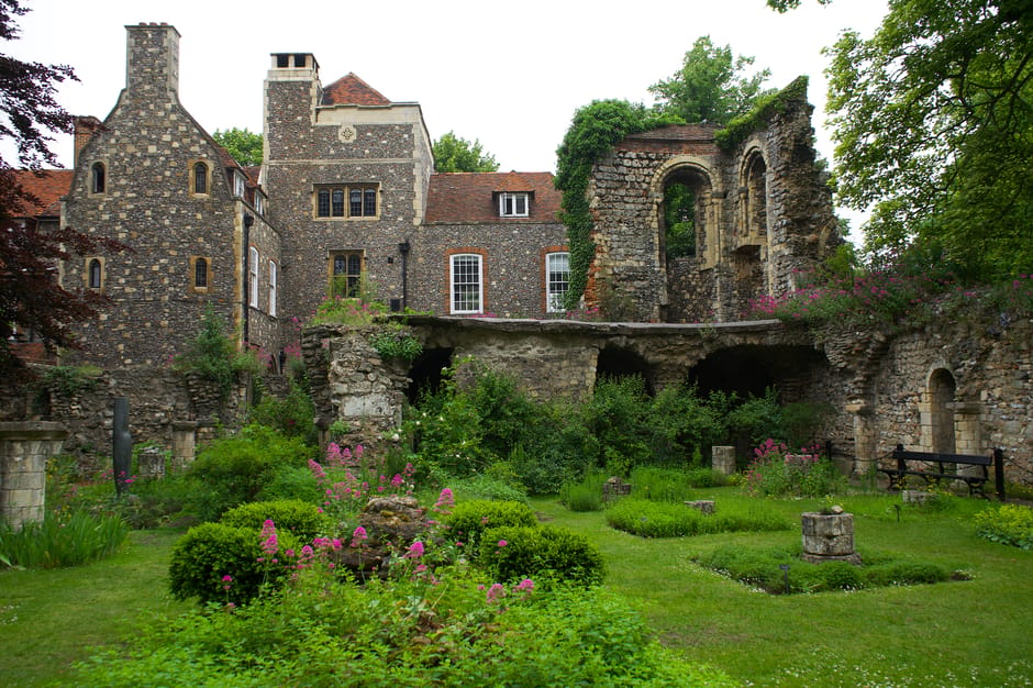 A scenic medieval-style garden featuring well-maintained green lawns and a variety of lush plants. In the foreground, there is a stone pedestal with a flat top, surrounded by flowerbeds with small pur